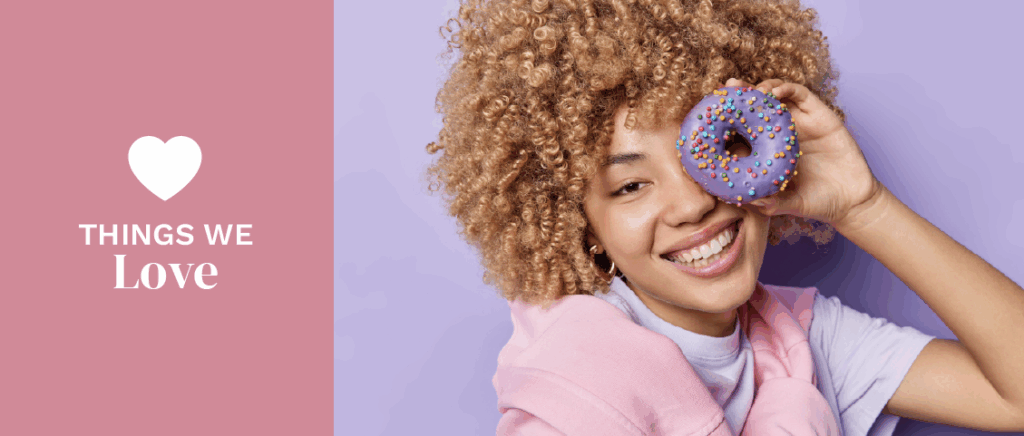 Glad curly haired woman covers eye with an appetizing glazed doughnut smiles happily